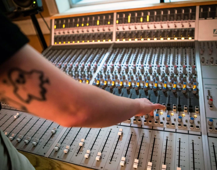 A close-up of a student's arm with a ghost tattoo reaching for controls on a large mixing desk, with glowing indicator lights. A close-up of a student's arm with a ghost tattoo reaching for controls on a large mixing desk, with glowing indicator lights.